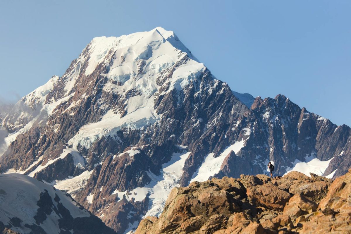 A lone hiker stands near Aoraki/Mount Cook in New Zealand under a clear sky.