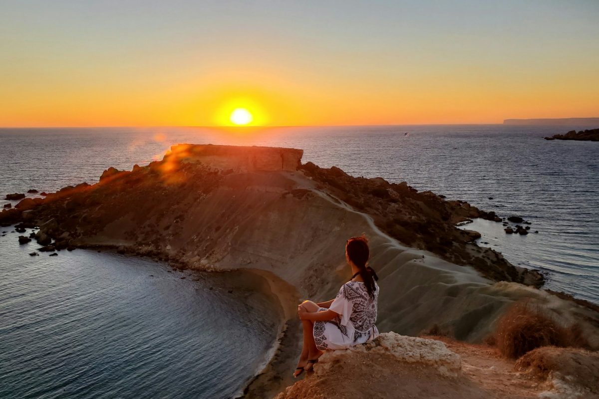 woman in white shirt standing on brown rock near body of water during sunset